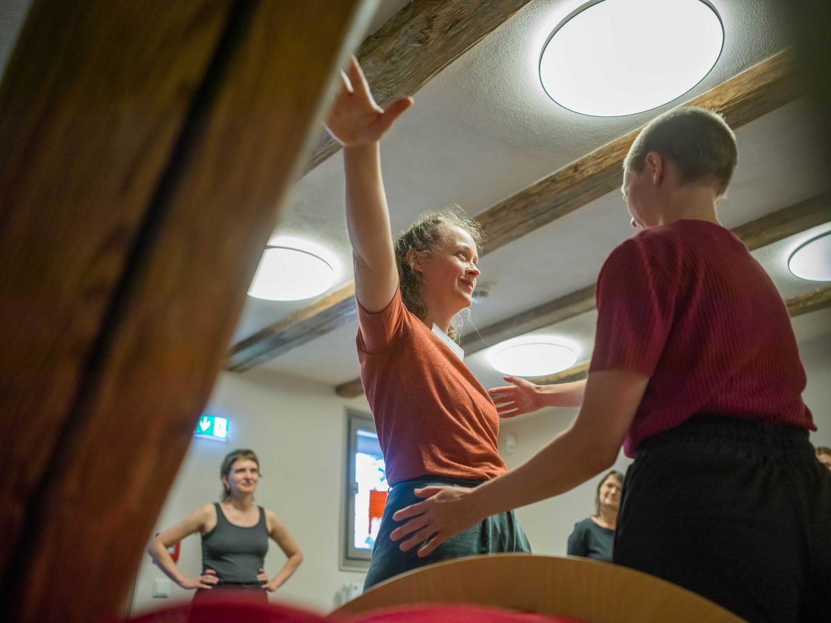 Man sieht zwei Frauen. Die eine Frau breitet die Arme aus. Die zweite Frau steht ihr gegenüber und hat ihre Arme auf Hüfthöhe ausgebreitet.