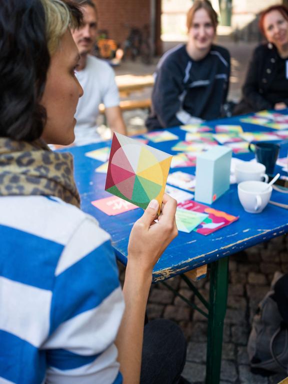 Man sieht einen Tisch an dem Menschen sitzen. Die Frau im Vordergrund hält eine Karte in der Hand.