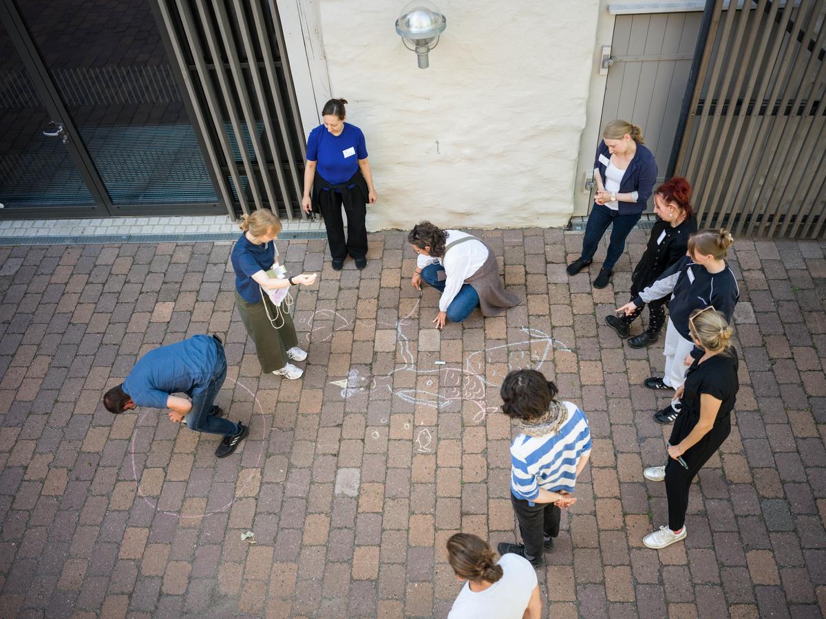 Das Bild zeigt eine Gruppe Menschen von oben, wobei zwei eine Kreidezeichnung anfertigen. Acht Menschen stehen im Kreis drumherum und schauen zu. 